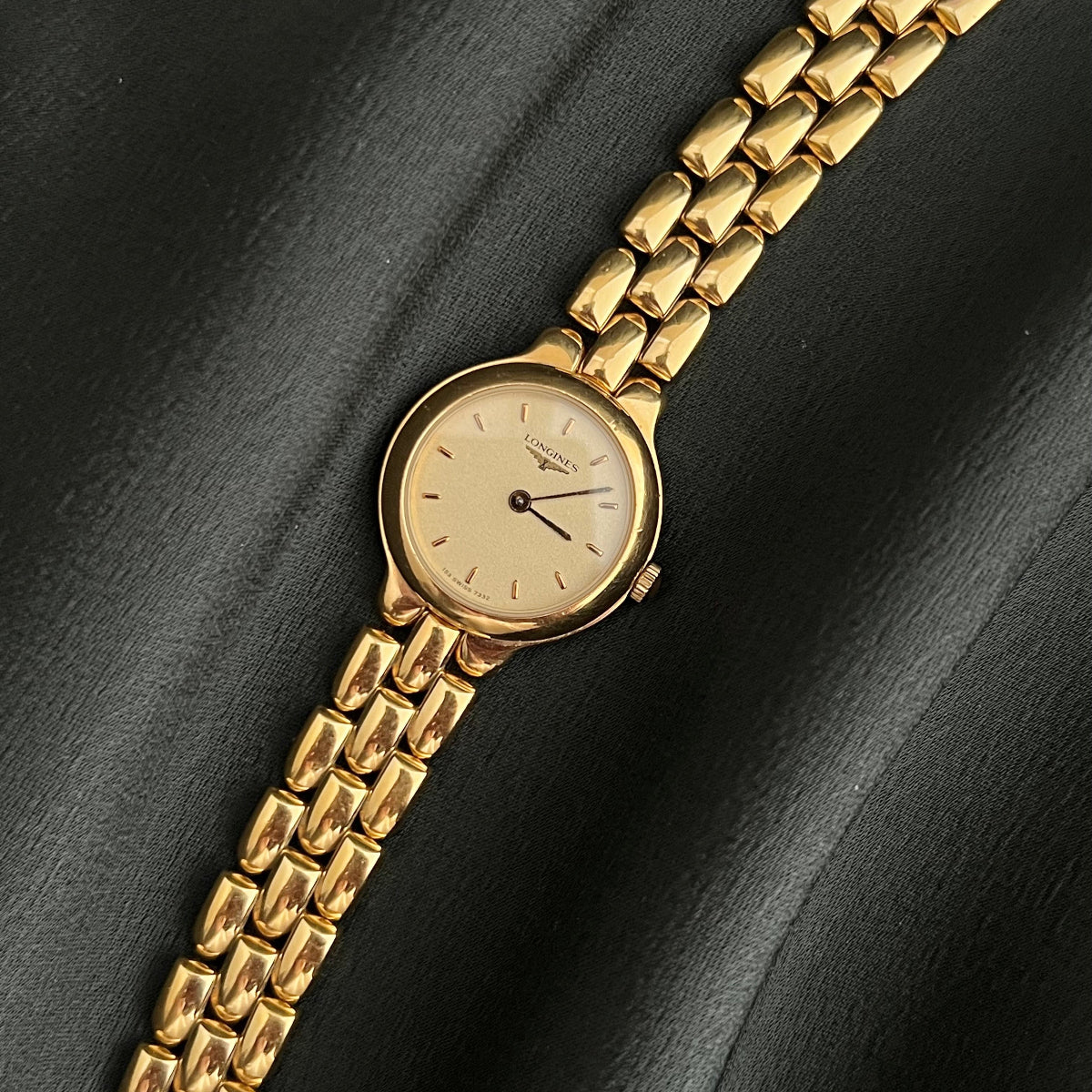 A vintage-style gold-tone wristwatch with a matching gold link bracelet, resting diagonally on a dark, draped satin fabric background.