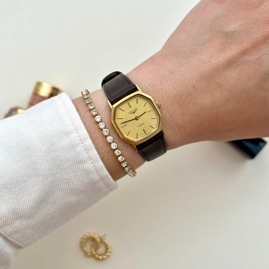 Hand wearing a gold Longines Women's watch with a black strap on a white background