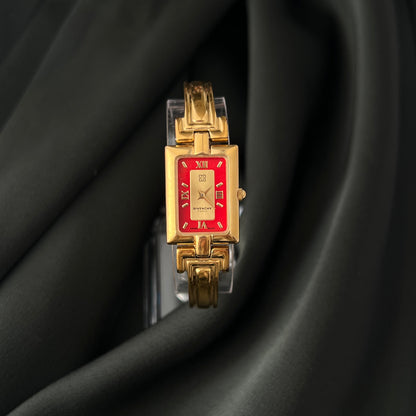 A gold-tone rectangular watch with a red dial and red Roman numerals, displayed on a black satin fabric background.