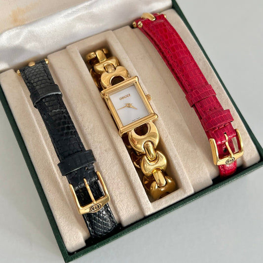 Gucci wristwatches presented in a dark green box with cream lining: a black textured strap watch, a gold-tone chain watch with a white dial, and a bright red textured strap watch.