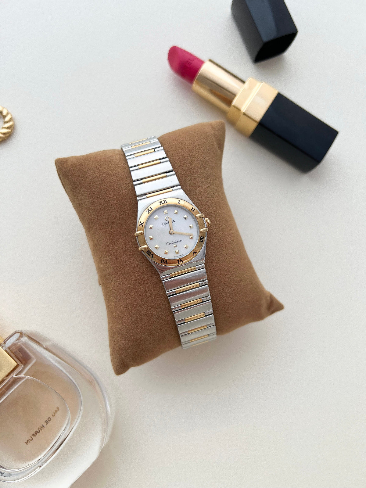 Omega Constellation Two Tone watch on a brown cushion with lipstick and perfume bottle in the background