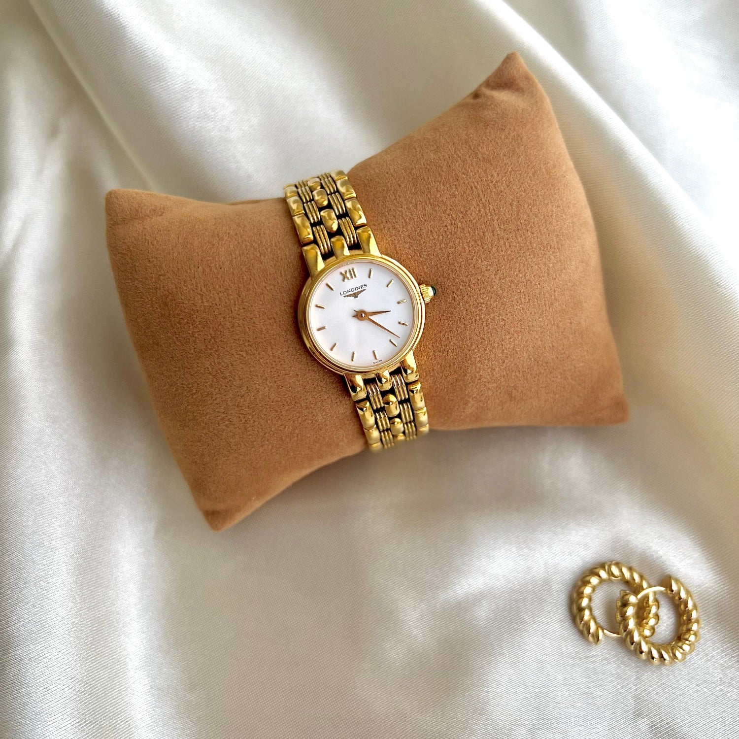 Gold Longines Women's watch with a green stone and earrings on a beige pillow with a white fabric background
