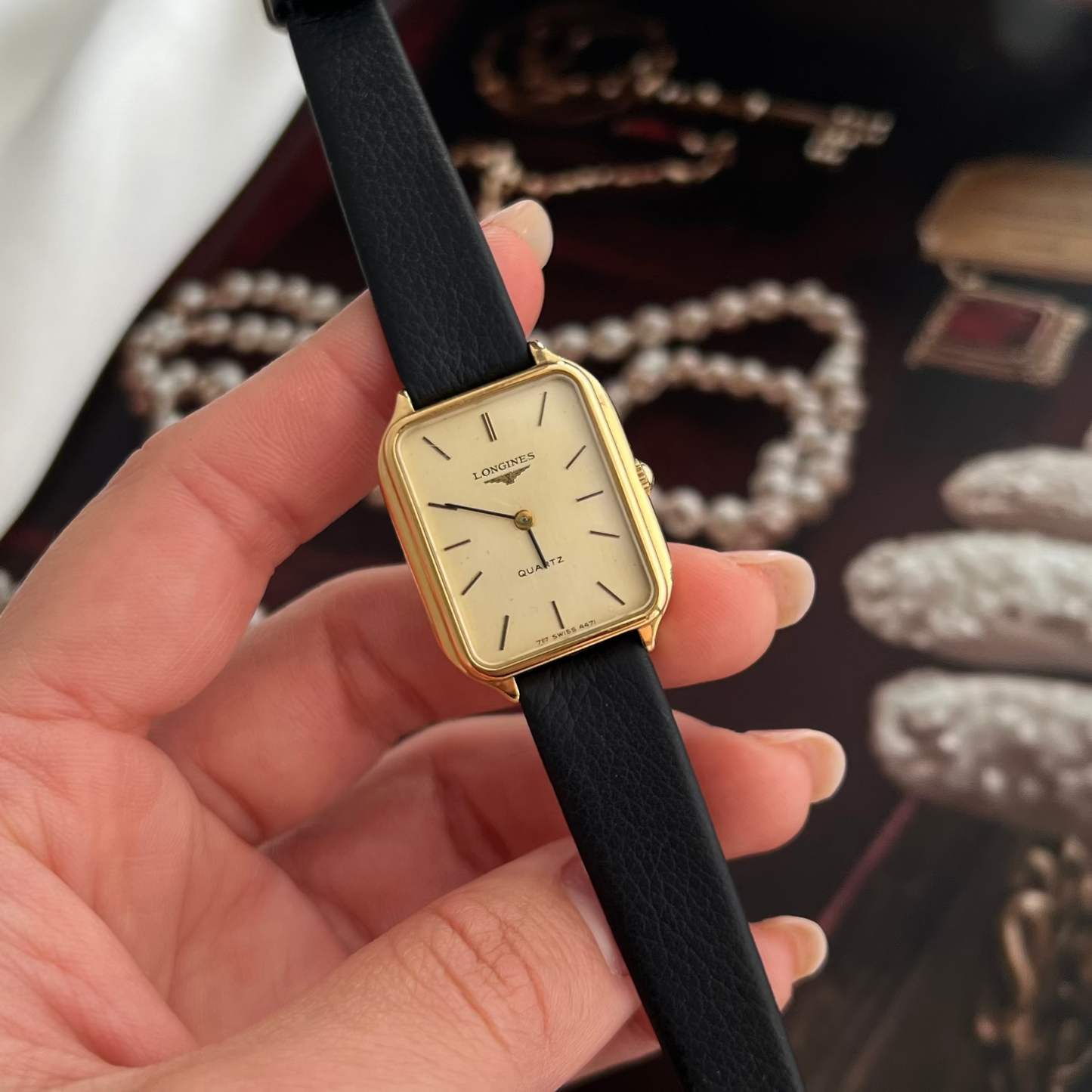 Gold Longines watch with black strap held in a hand against a jewelry display background