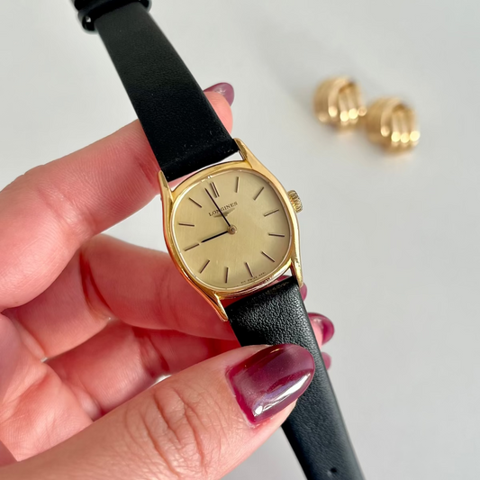 Hand holding a gold Longines watch with black strap against a light gray background