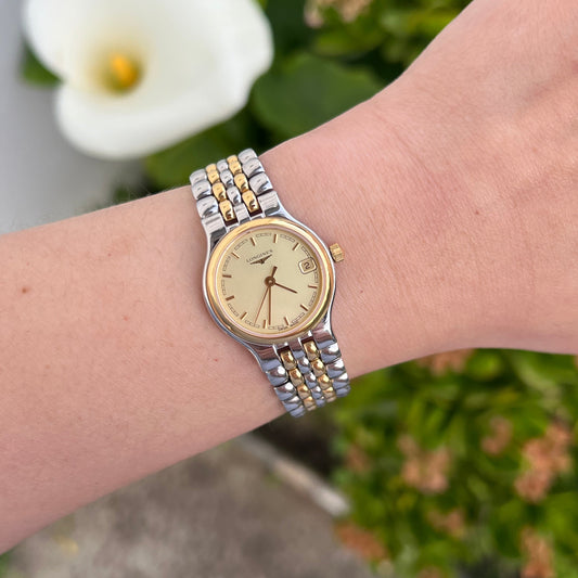 A two-tone silver and gold wristwatch with a gold dial, worn on a person's wrist against a blurred background of green foliage and a white flower.