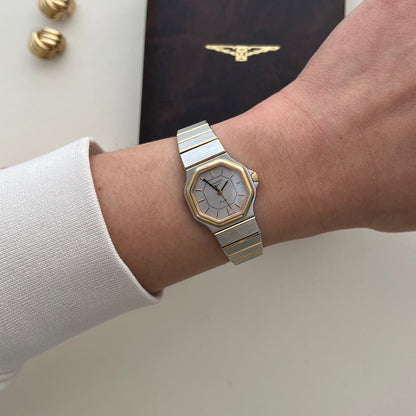 A Longines silver-toned metal link bracelet watch with an octagonal bezel and white dial, worn on a person's wrist. A dark brown watch box with a gold logo and two gold cufflinks are placed on a light beige surface in the background.