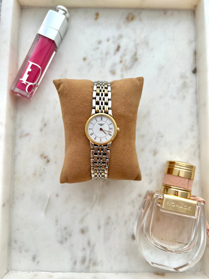 Gold Longines watch on a brown cushion with a pink lip gloss and perfume bottle on a marble surface