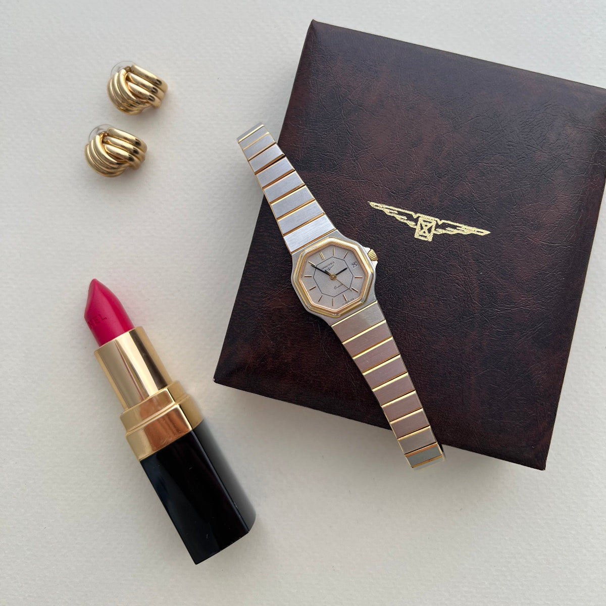 A Longines luxury women's watch with a gold-toned octagonal case and a silver-and-gold striped metal bracelet, displayed on a dark brown textured box. The watch is styled in a flat-lay composition alongside a pink lipstick and a pair of gold cufflinks on a light gray background.