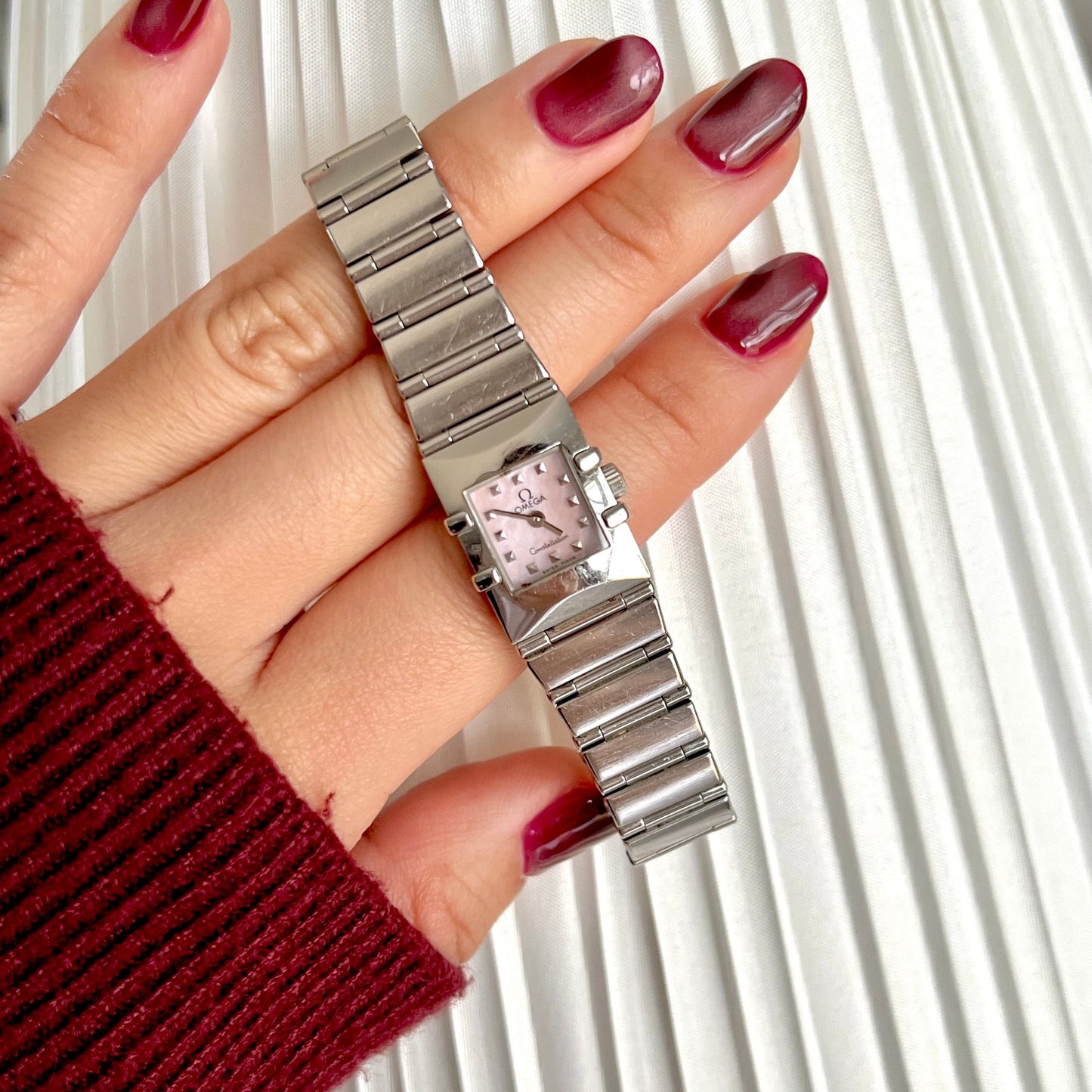 Silver Omega Constellation Women's watch with a pink face held by a hand with red nail polish against a white background