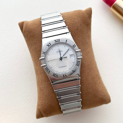 Omega Constellation Silver watch on a brown cushion with a blurred lipstick in the background