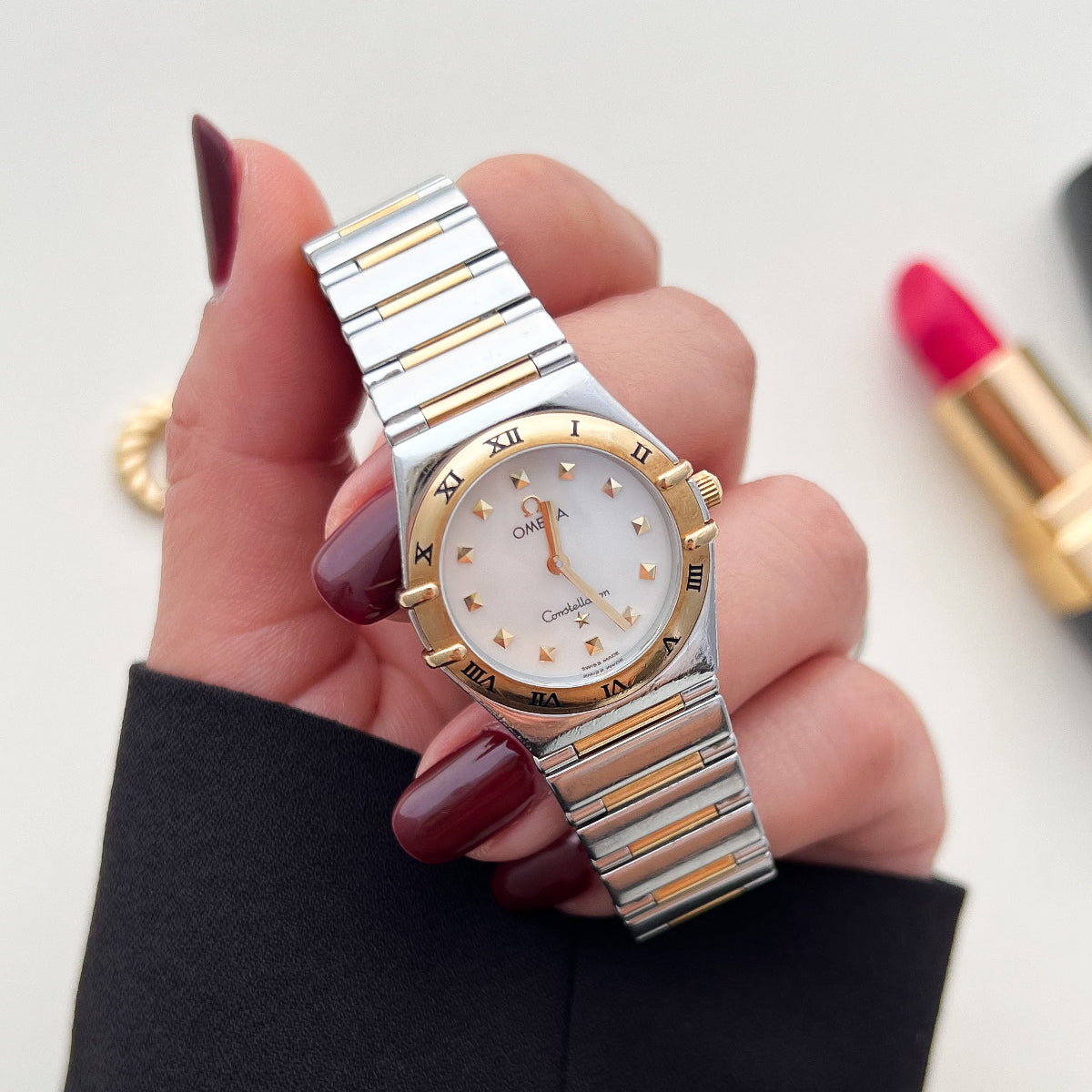 Omega Constellation Women's Watch in Two ToneHand holding a silver and gold watch with a blurred lipstick and ring in the background