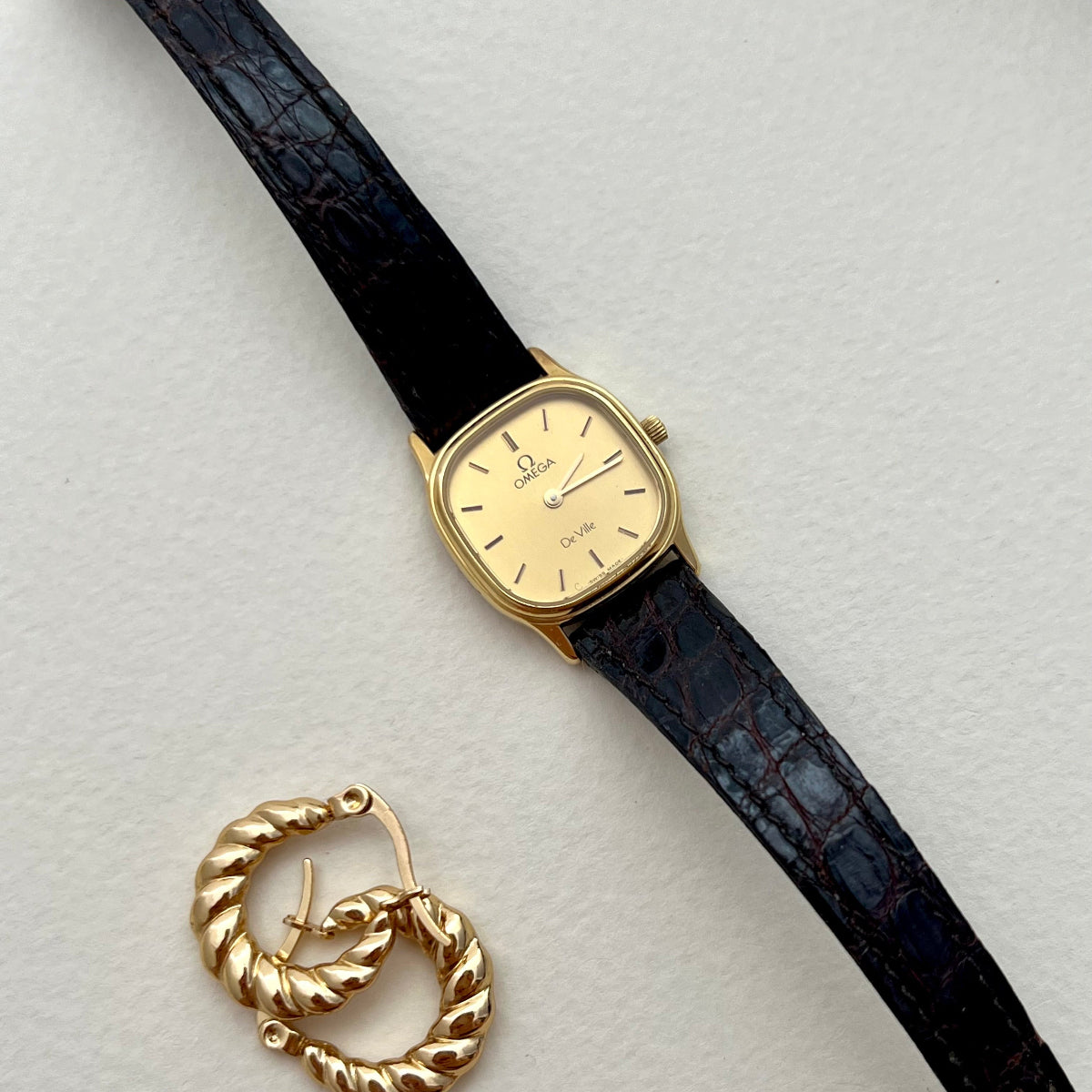 Omega Gold Luxurious Women's watch with black strap and gold hoop earring on a light gray background