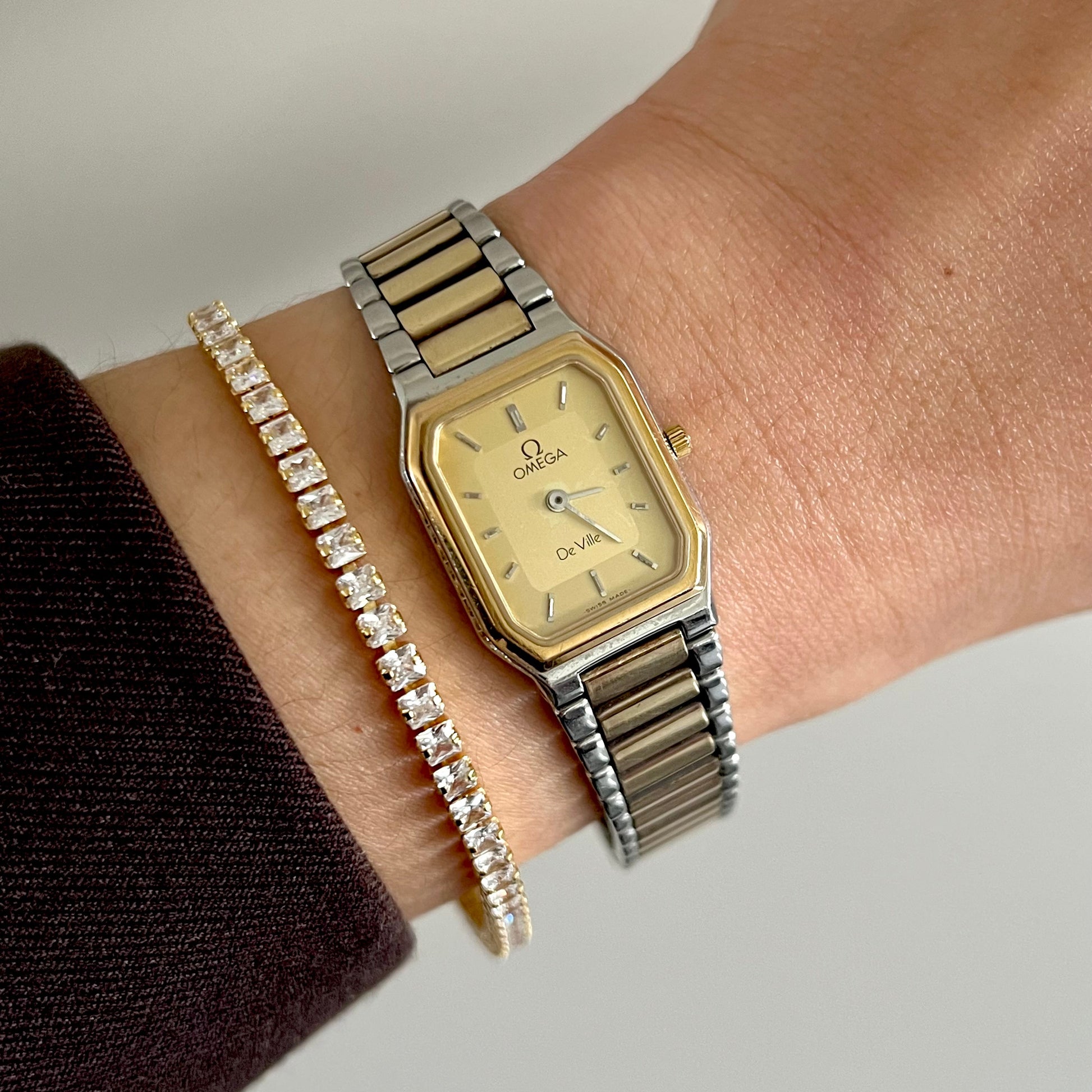 A close-up of a person's wrist wearing an Omega Quartz watch with a two-tone gold and silver metal bracelet, alongside a gold tennis bracelet.