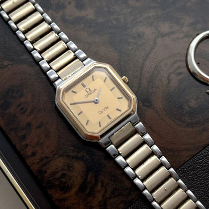An Omega Quartz wristwatch with a gold-toned dial and a two-tone silver and gold metal link bracelet, resting on a dark wood-grain surface.