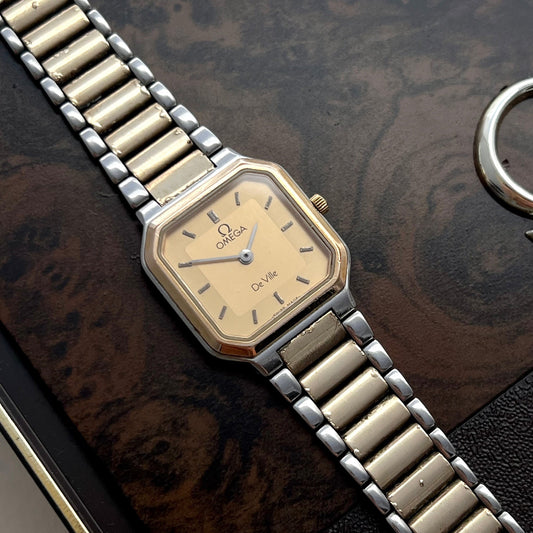 An Omega Quartz wristwatch with a gold-toned dial and a two-tone silver and gold metal link bracelet, resting on a dark wood-grain surface.