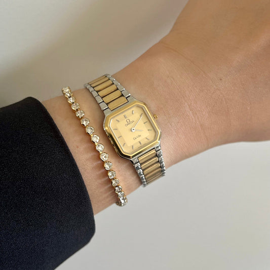 Gold Omega De Ville America watch and bracelet on a wrist against a neutral background