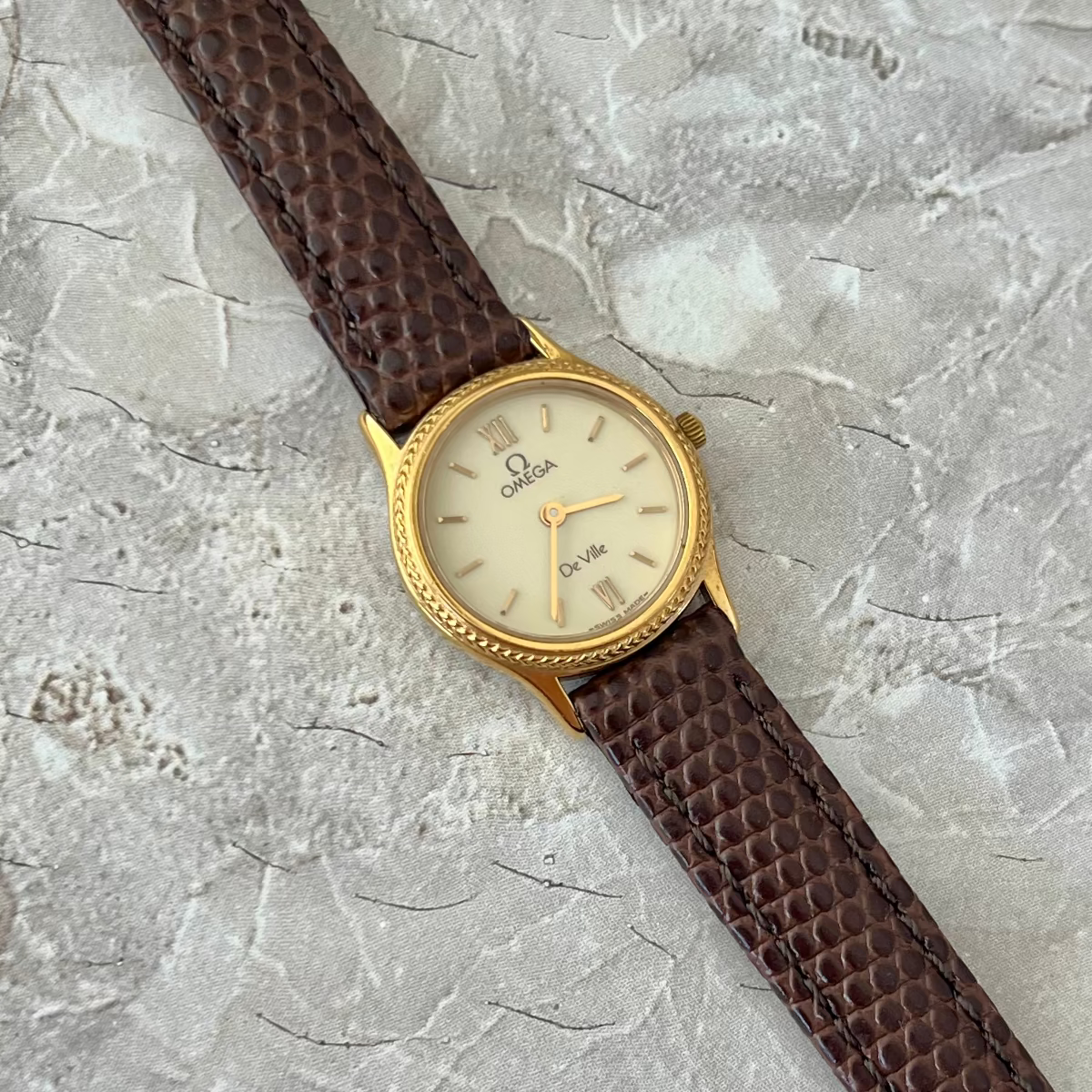 An Omega De Ville Ladies wristwatch with a textured brown strap and a gold-tone case, displayed diagonally on a light gray stone-textured background.