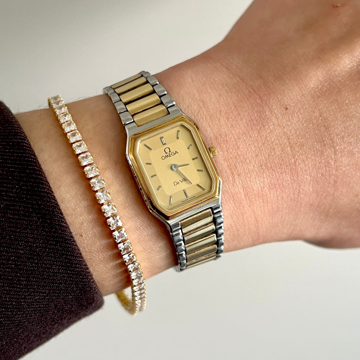 A gold and silver two-tone Omega De Ville Rectangular watch with a gold dial, worn on a person's wrist alongside a gold beaded bracelet.