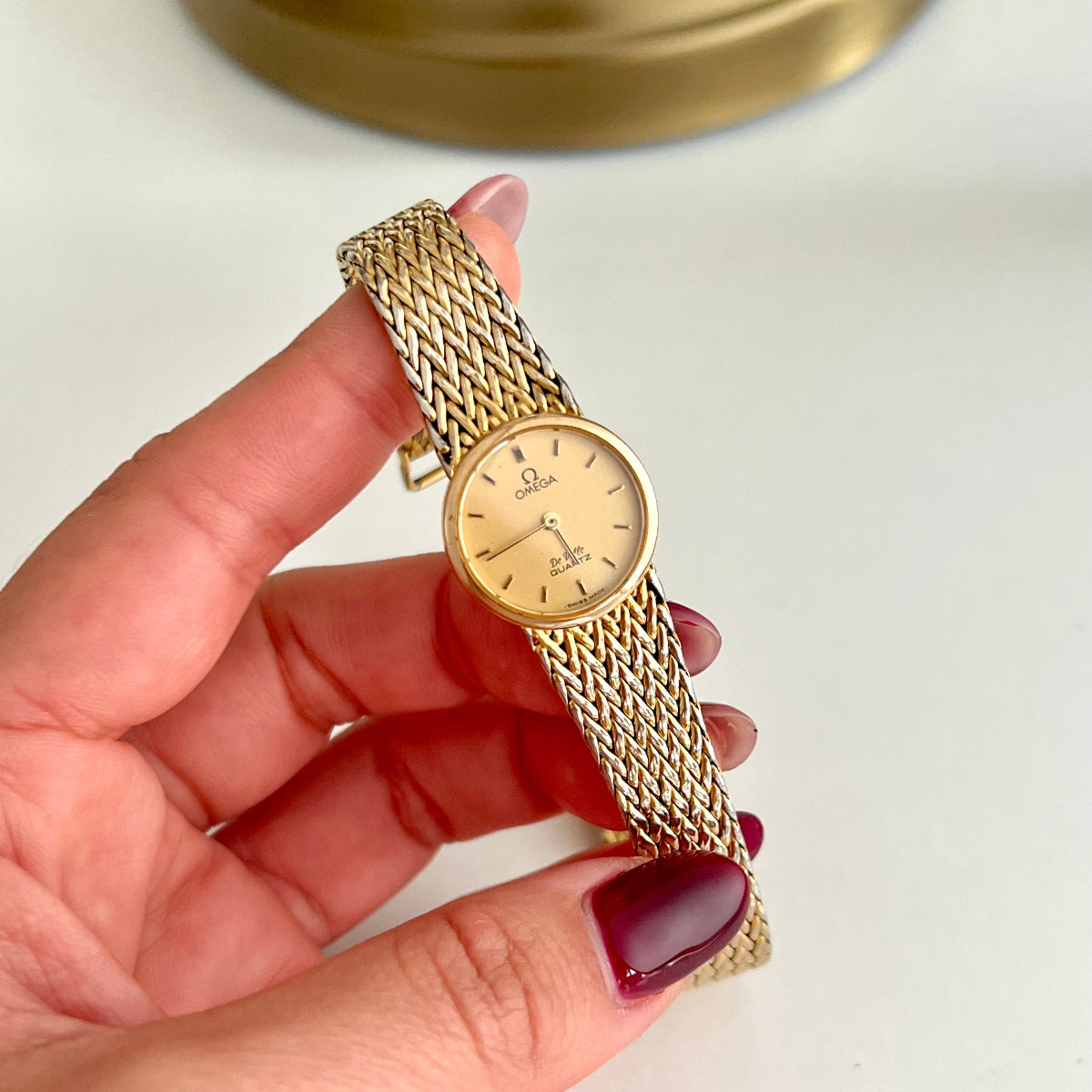Omega De Ville Gold watch held by a hand with red nail polish on a light background