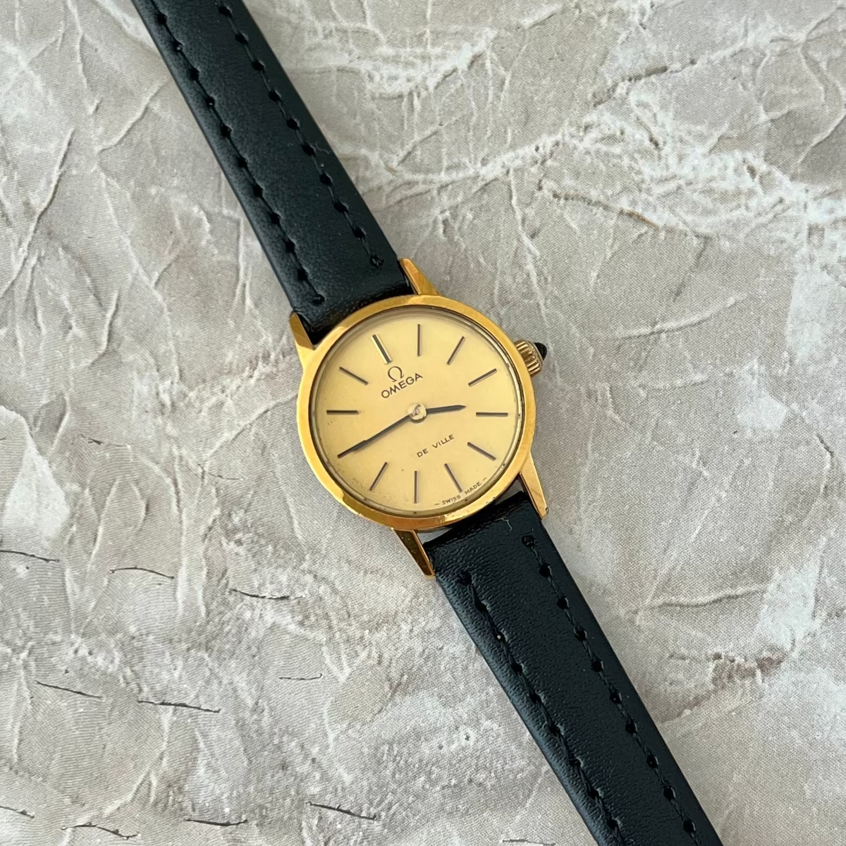 An Omega wristwatch with a gold-tone case and cream dial, paired with a black leather strap, laid diagonally on a textured light gray stone surface.