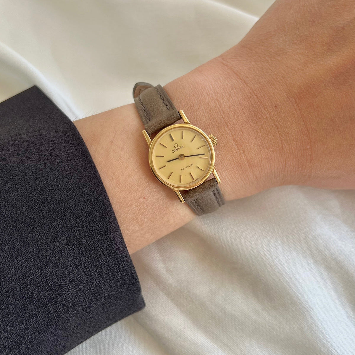 Gold Omega De Ville Women's watch with brown leather strap on a wrist, with earrings in the background.