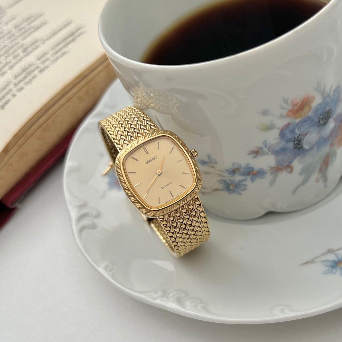 GSeiko Exceline gold watch on a saucer with a cup of coffee and book in the background