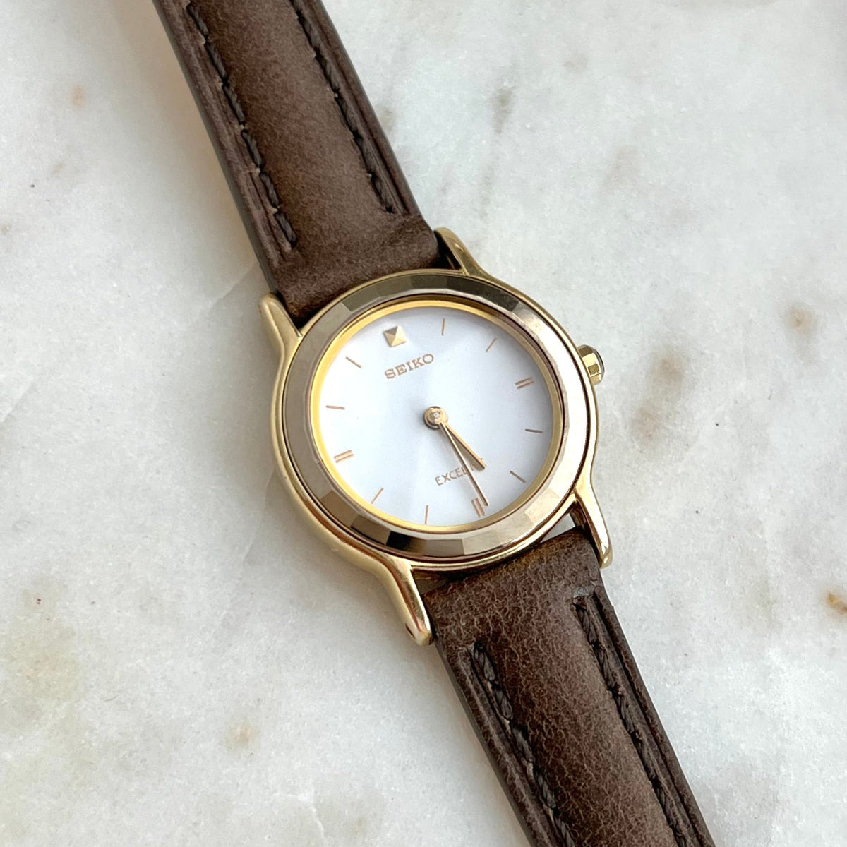 Seiko Exceline Women's Watch with Tungsten Carbide Faceted Bezel with brown leather strap and gold case on a marble surface