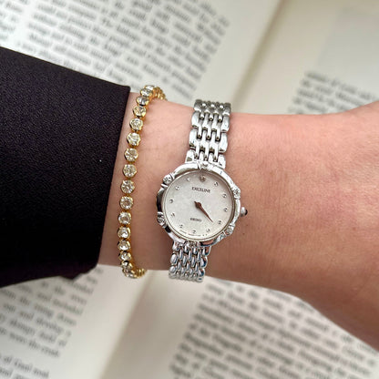 Silver Seiko women's watch on a wrist with a gold bracelet against an open book background