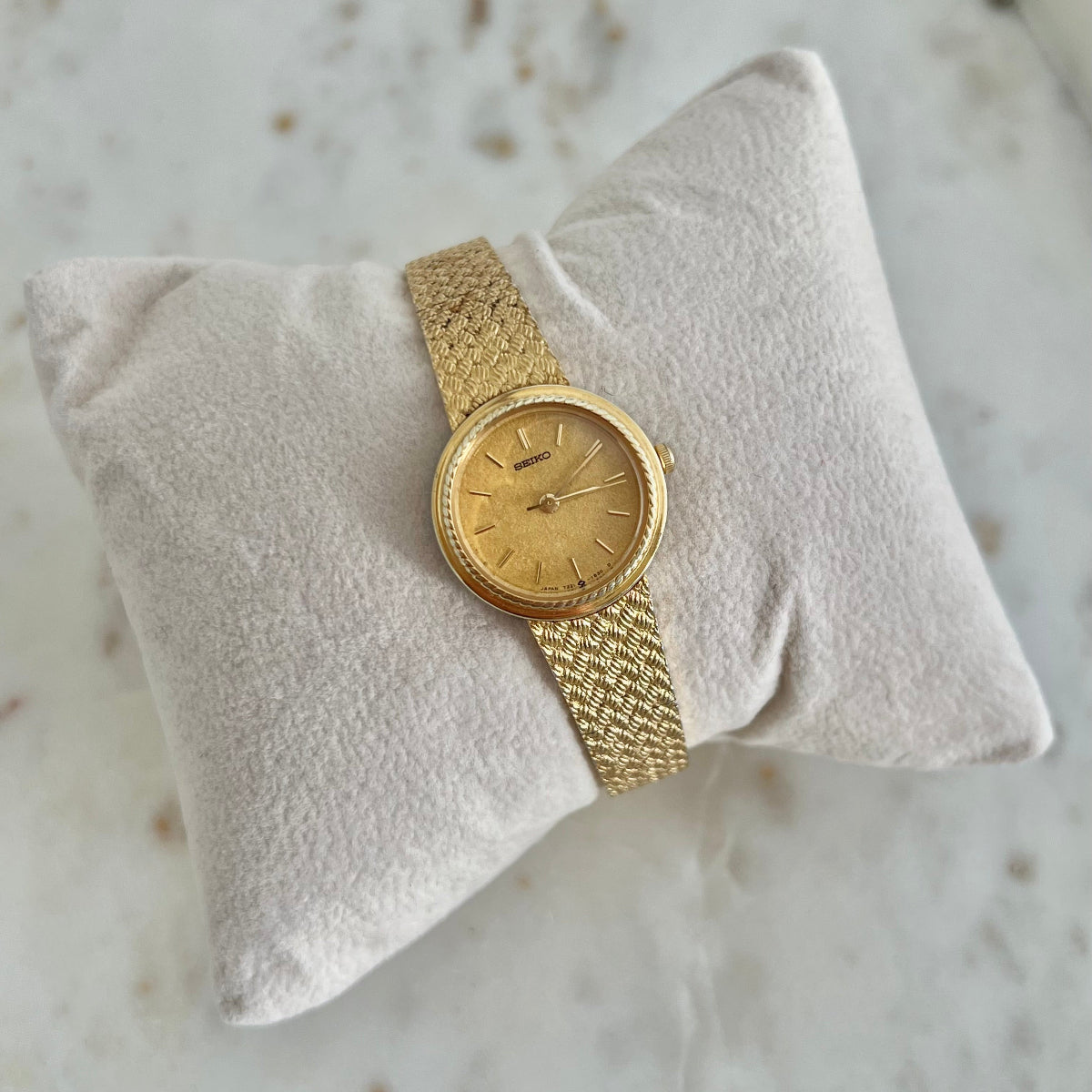 A vintage gold-tone wristwatch with a matching mesh metal band, displayed on a textured cream-colored pillow over a light marble surface.