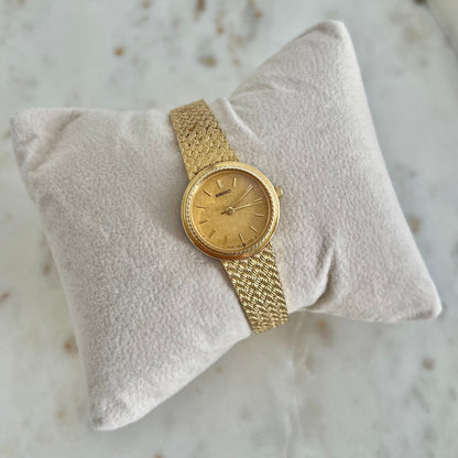 A vintage gold-tone wristwatch with a matching mesh metal band, displayed on a textured cream-colored pillow over a light marble surface.