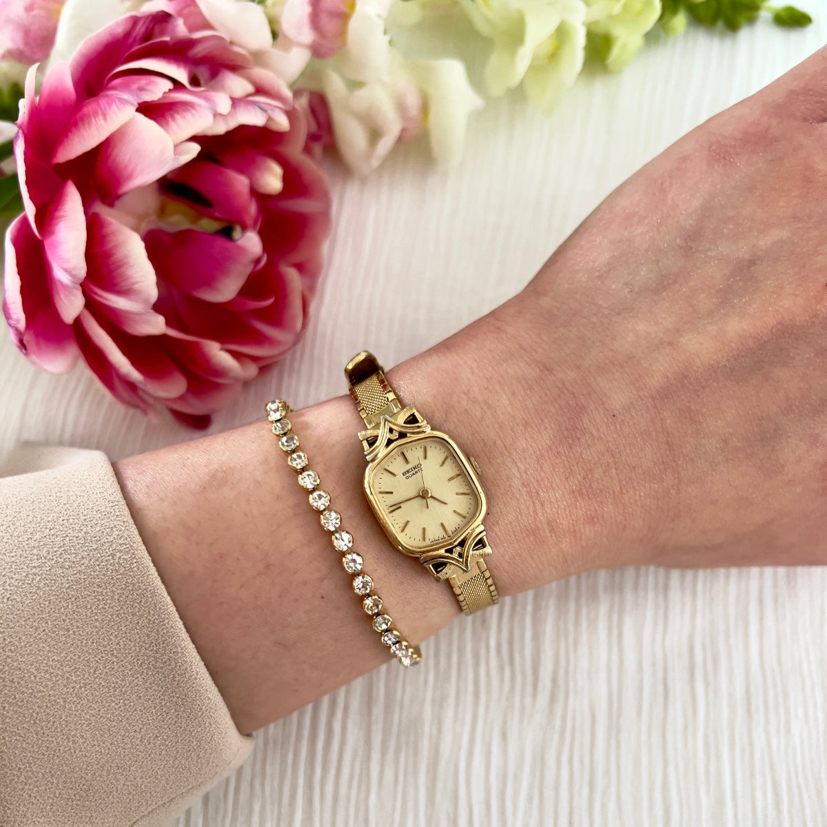 Seiko gold toned watch and bracelet on a wrist with pink flowers in the background