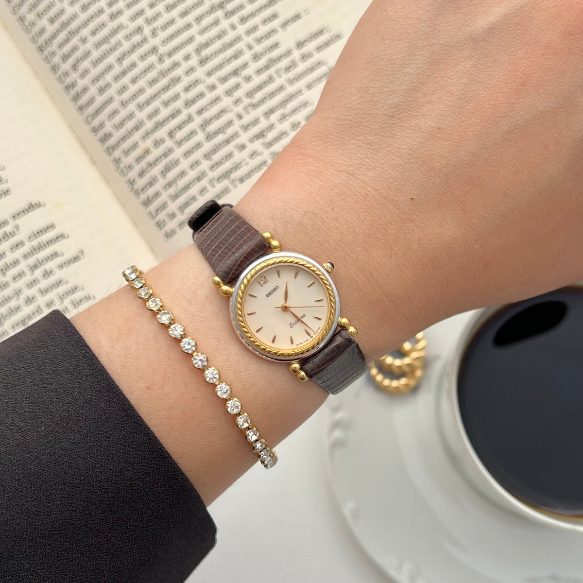 Hand wearing a Seiko two tone watch with a brown strap on a surface with an open book and a cup of coffee.