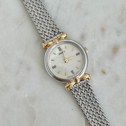A vintage-style wristwatch with a silver mesh strap, round silver case, and gold-tone bow details on either side of the white dial, photographed on a light marble surface.