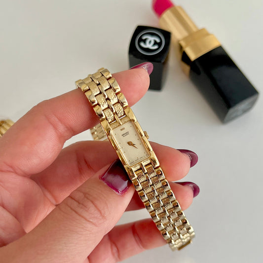 Gold Seiko women's watch held by a hand with a blurred background of lipstick and nail polish.