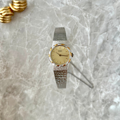 Editorial flat lay of a late century Seiko Women’s watch featuring a champagne dial, twisted gold bezel accents and a wide crosshatched silver bracelet, styled with gold earrings on softly draped white satin.