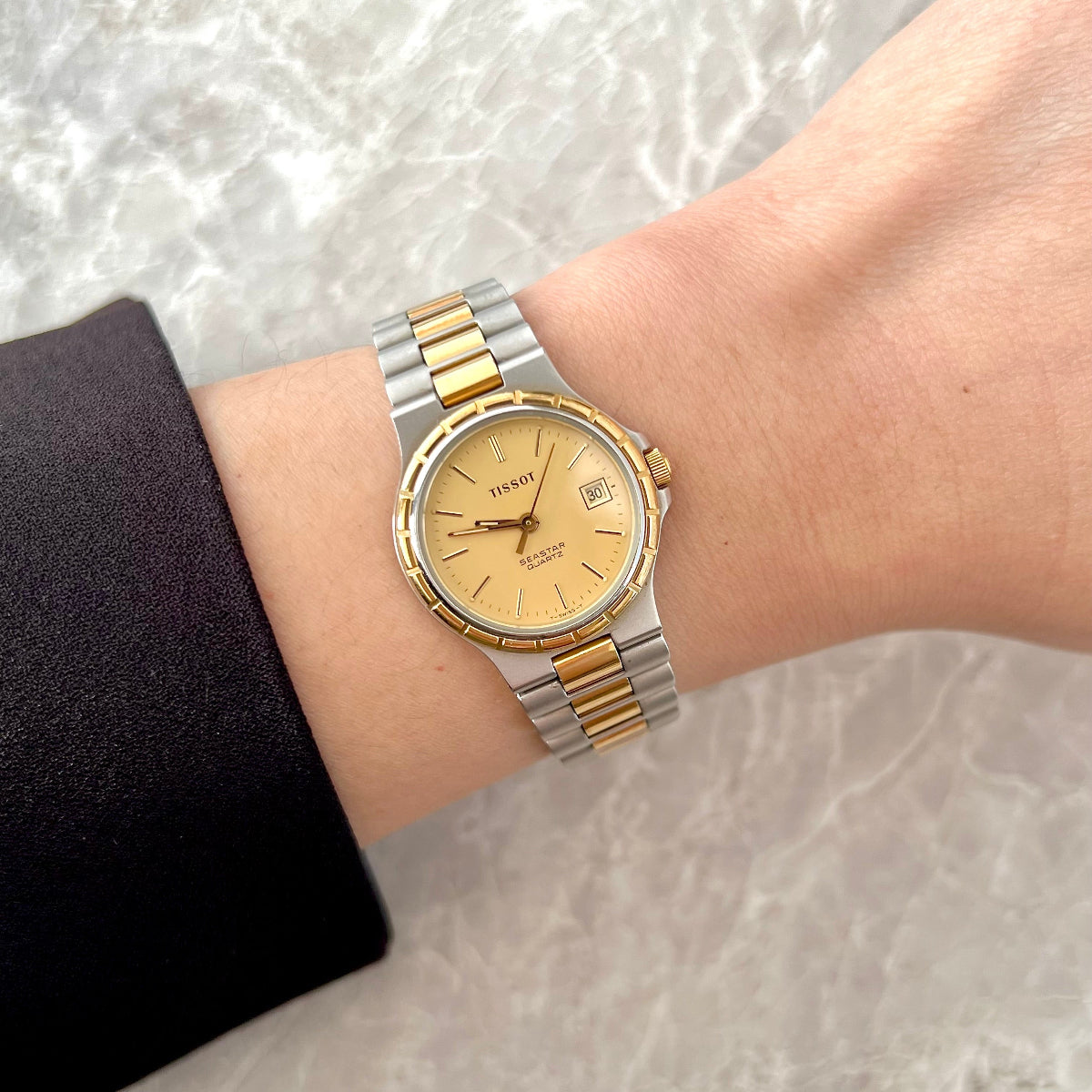 A two-tone gold and silver wristwatch with a gold dial, worn on a person's wrist against a light marble background.