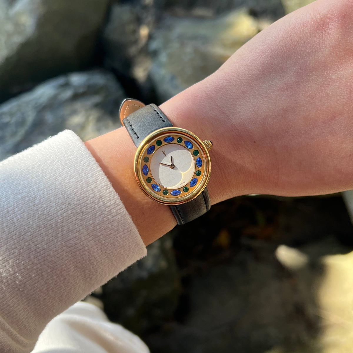 A close-up of a wrist wearing a gold-tone YSL watch with a white dial, blue and yellow stone-set bezel, and a light blue leather strap, shown outdoors against a blurred rocky background.