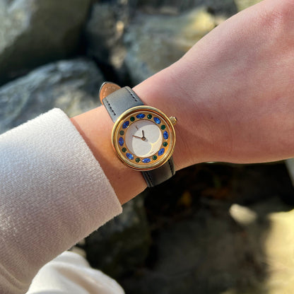 A close-up of a wrist wearing a gold-tone YSL watch with a white dial, blue and yellow stone-set bezel, and a light blue leather strap, shown outdoors against a blurred rocky background.