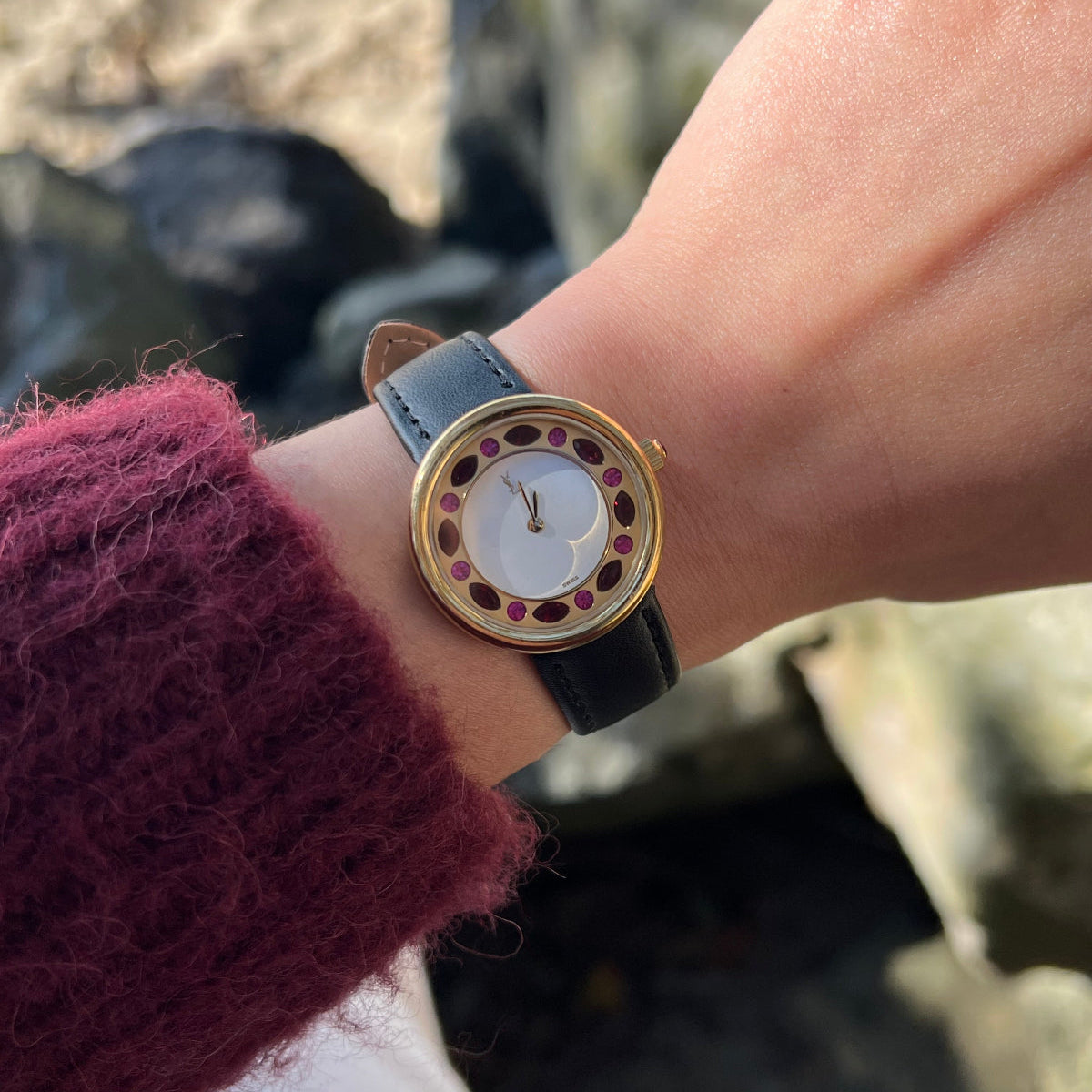 A close-up of a wrist wearing a round YSL Rubies watch with a gold-tone case, white dial, and dark strap, shown against an outdoor blurred background with a person's hand and sleeve visible.