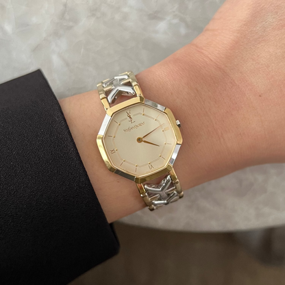 A gold-tone and silver-tone two-tone YSL watch with a cream dial, worn on a person's wrist. The watch has a unique geometric octagonal case and a matching link bracelet with silver-toned star-like accents.