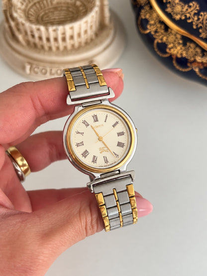 Burberrys cream dial with outlined Roman numerals, gold tone hands and equestrian knight emblem, front view held in a hand.