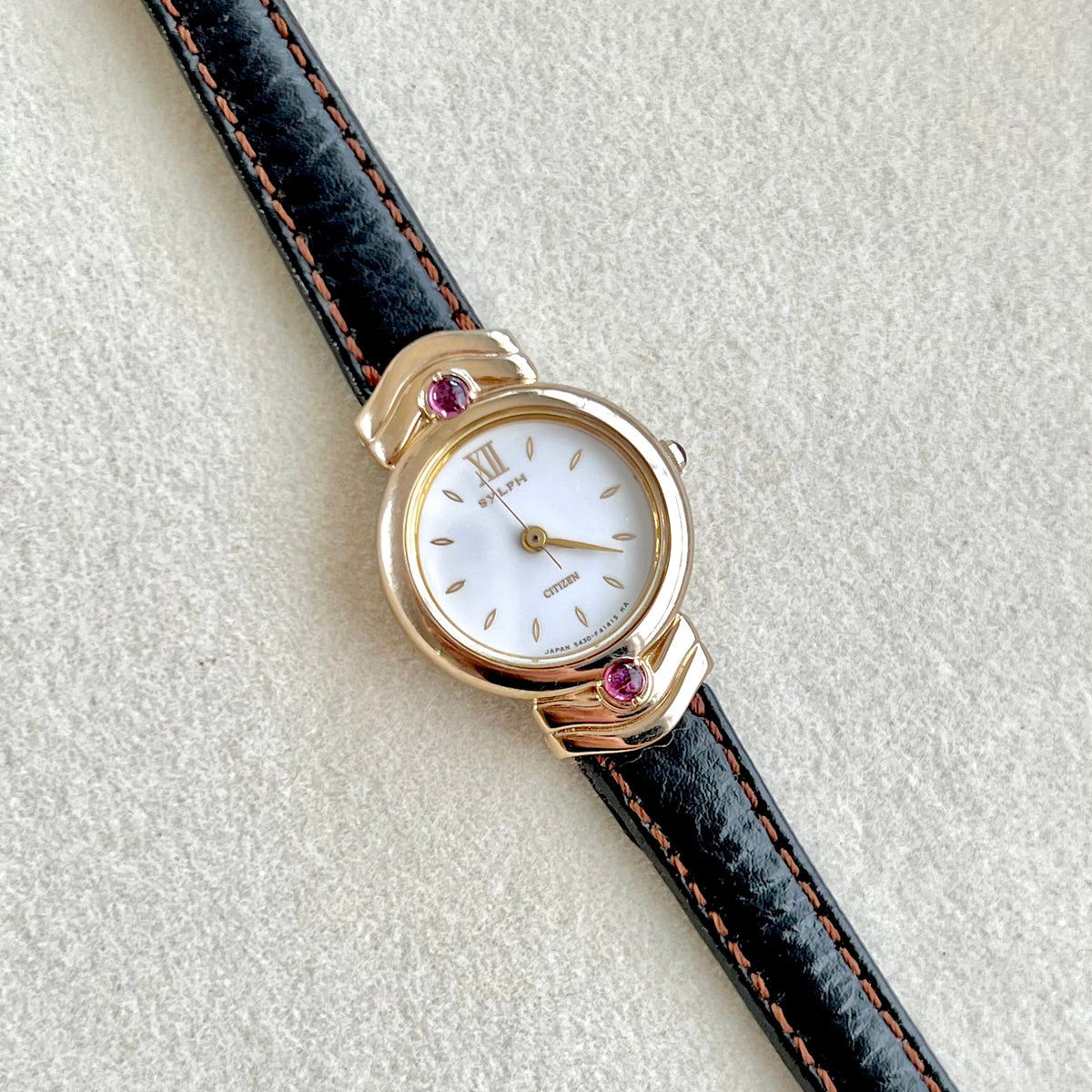 Citizen women’s watch with a gold-tone case set with purple gemstones, white dial, and leather strap with red stitching. (Citizen Sylph model).
