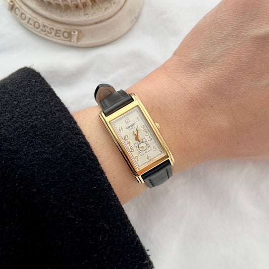 Gruen women’s watch worn on wrist featuring a gold tone rectangular case, Art Deco inspired dial, small seconds display and black leather strap.
