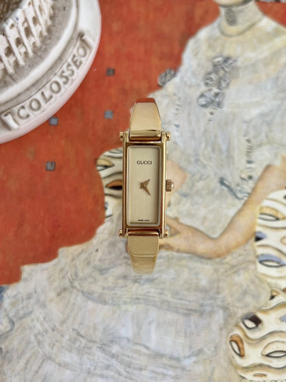 Gucci 1500 women’s gold tone horsebit bangle watch with a narrow rectangular champagne dial, photographed from the front