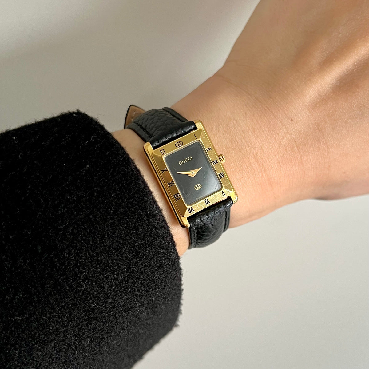 Gucci women’s watch with black dial, Roman numeral bezel, gold tone rectangular case and black leather strap worn on wrist