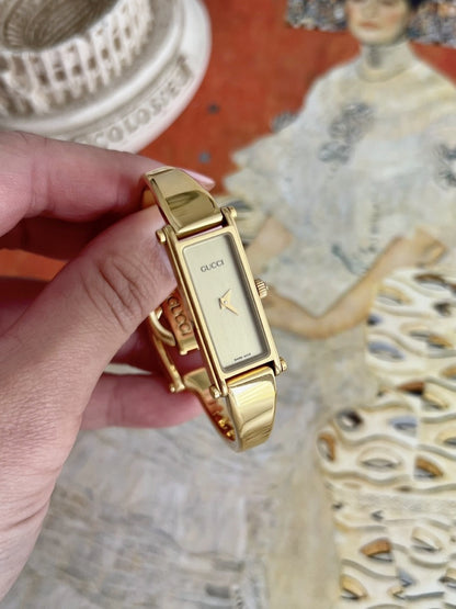 Gucci women’s rectangular champagne dial gold tone bangle watch held to show its case and bracelet profile