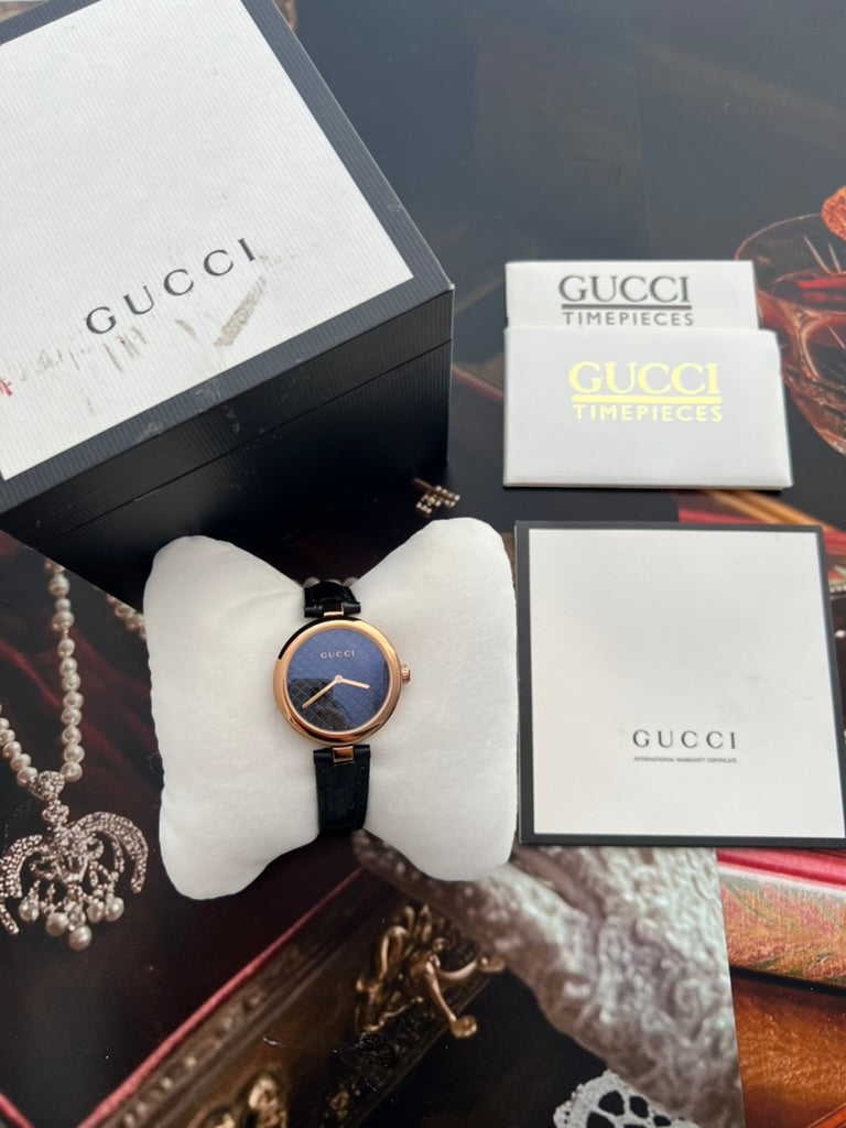 Front styling view of the Gucci Diamantissima round rose gold tone watch and black leather strap on a cushion.