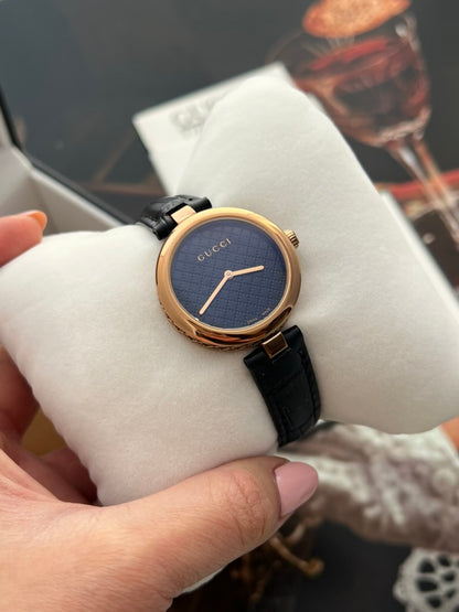 Gucci Diamantissima women’s watch held on a cushion, showing its dark patterned dial and slim rose gold tone hands.