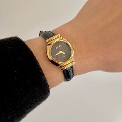 Gucci women’s watch with black oval dial, gold tone case, baguette inspired lugs and black leather strap worn on wrist