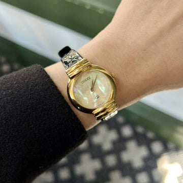 Gucci women’s watch on wrist with an iridescent mother of pearl dial, rounded gold tone case, ribbed lugs and black leather strap